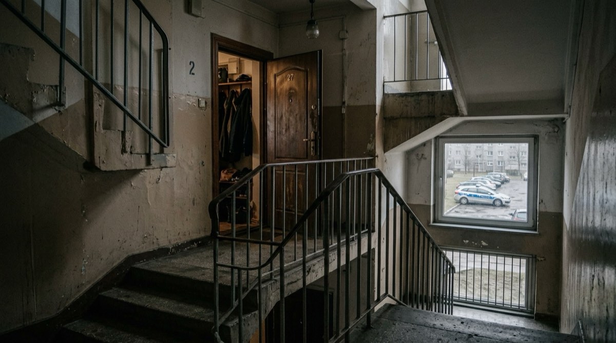 Klatka schodowa w bloku, uchylone drzwi mieszkania, w tle radiowóz na parkingu